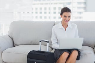 Happy businesswoman sitting on the couch using laptop with her suitcase beside her.jpeg